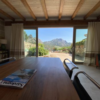 Contattaci per informazioni e prenotazioni. Prenota ora la tua vacanza esclusiva.Vista panoramica dal soggiorno di Casa Graniti, con colline verdi e il borgo di San Pantaleo sullo sfondo, immerso nella natura sarda.